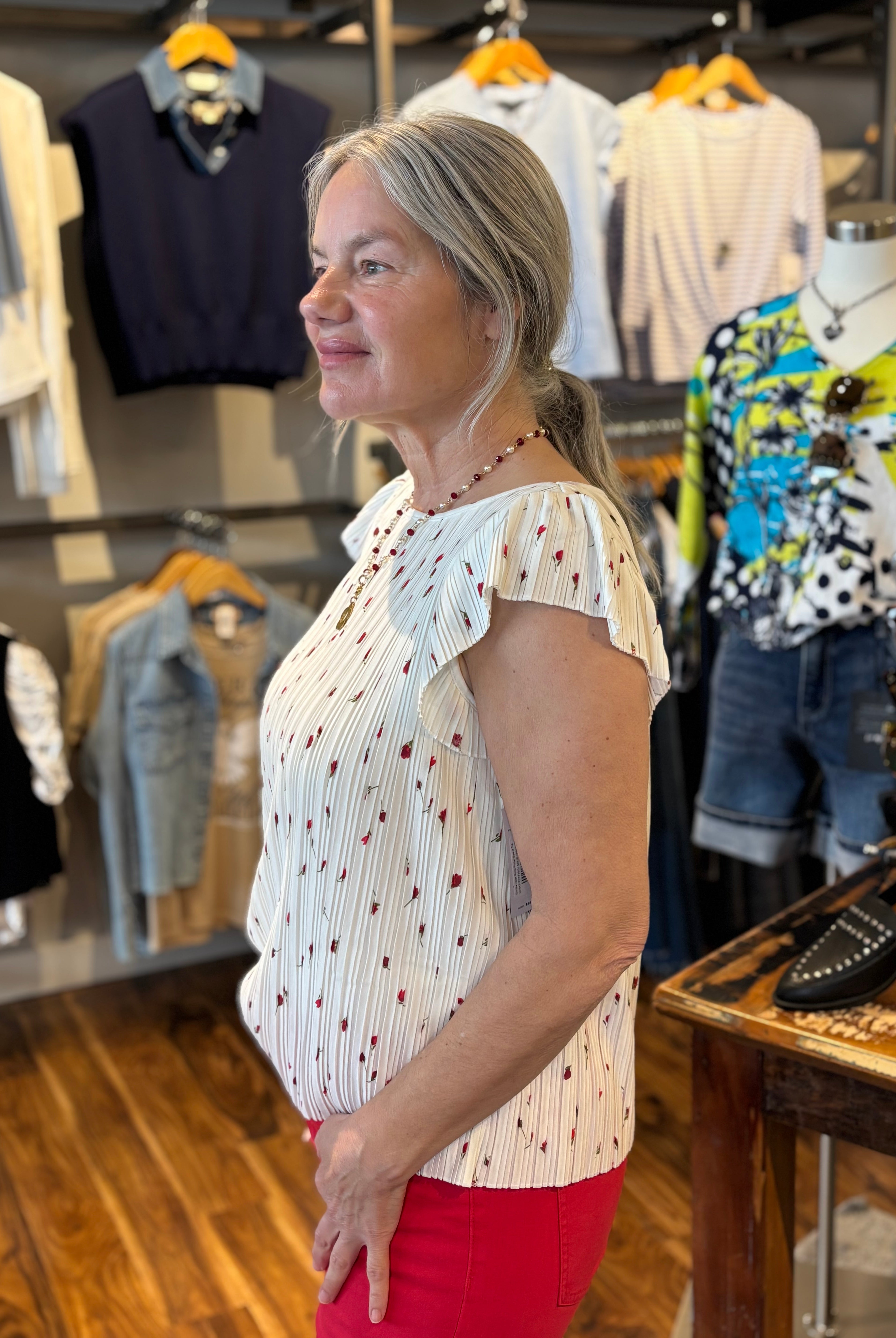Woman standing in a clothing store wearing a white flutter sleeve top with small red flowers and green stems