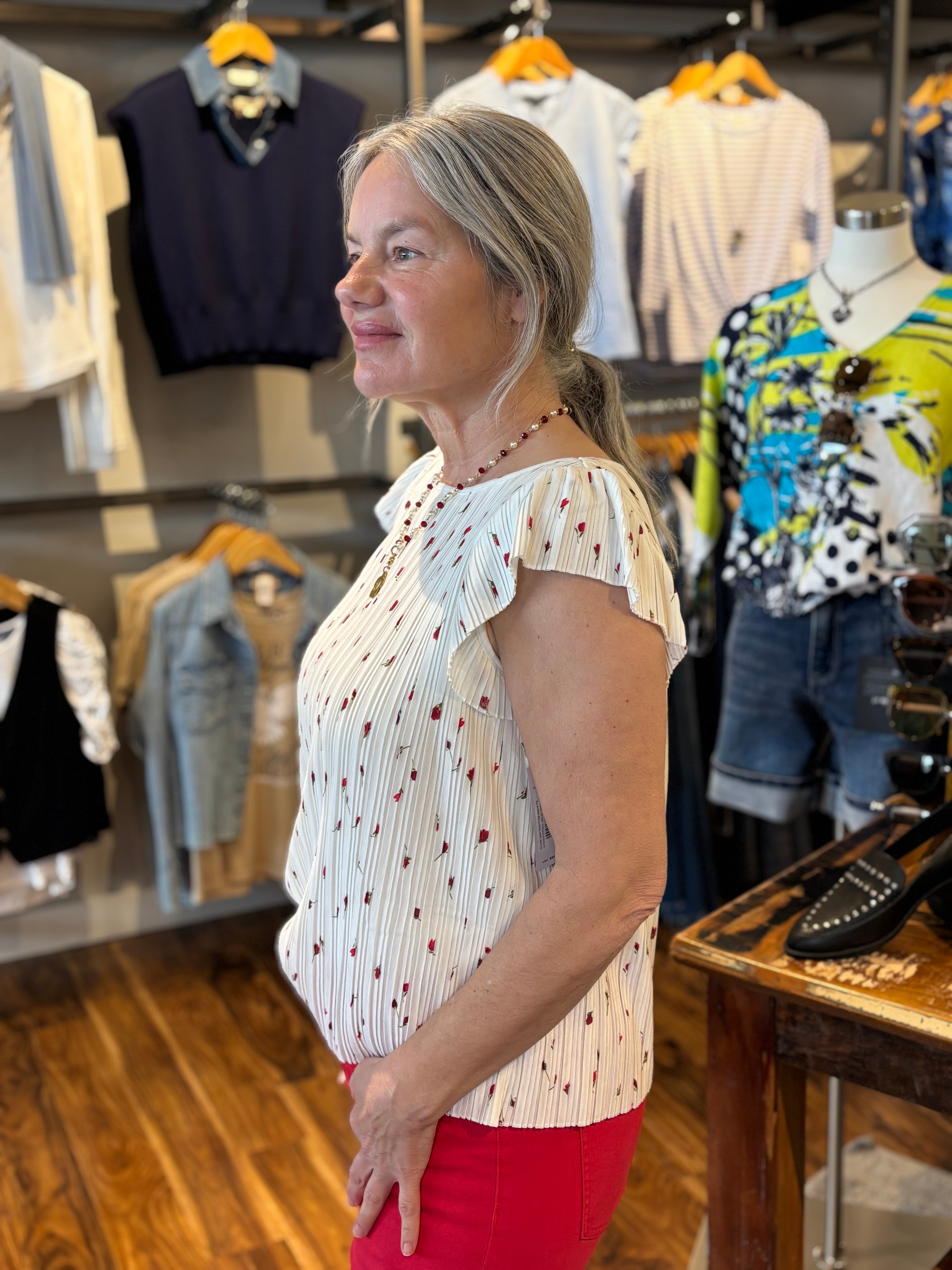 Woman standing in a clothing store wearing a white flutter sleeve top with small red flowers and green stems